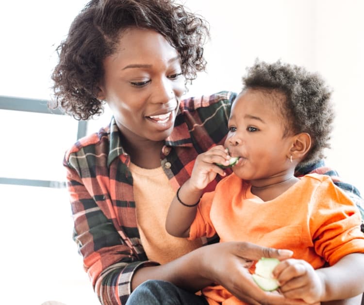 Toddler happily eating food while smiling mom holds them