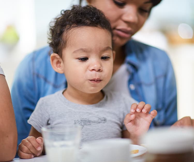 Mom helping toddler boy with his food at the dinner table