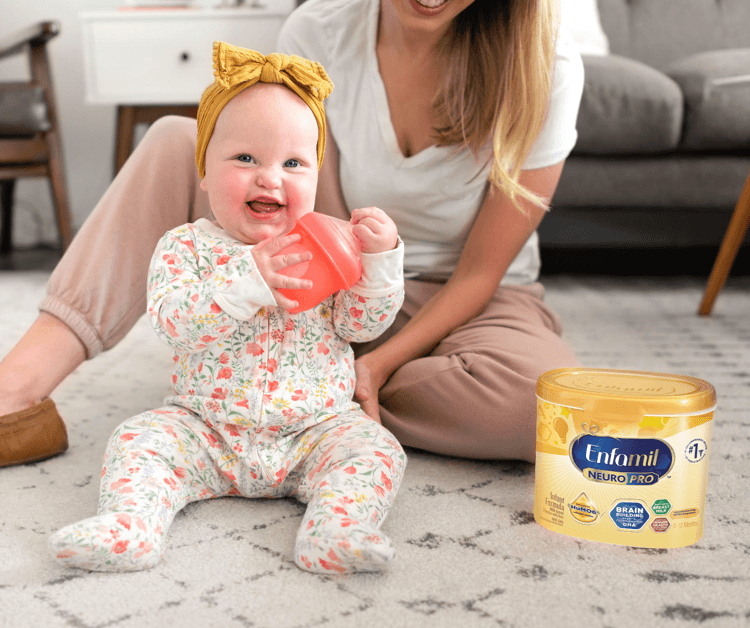 Baby smiling and holding bottle while sitting next to Mom on the floor
