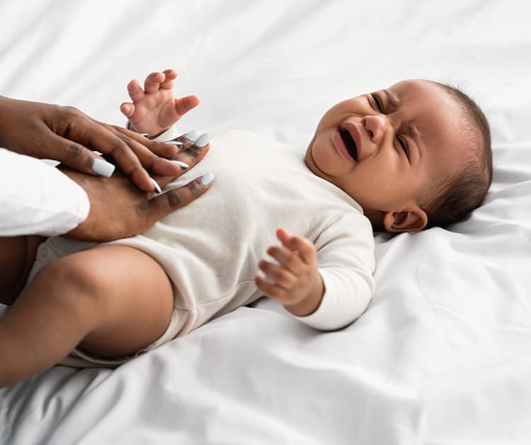 A baby lying on a white bed, crying, while an adult gently places their hands on the baby's stomach for comfort.
