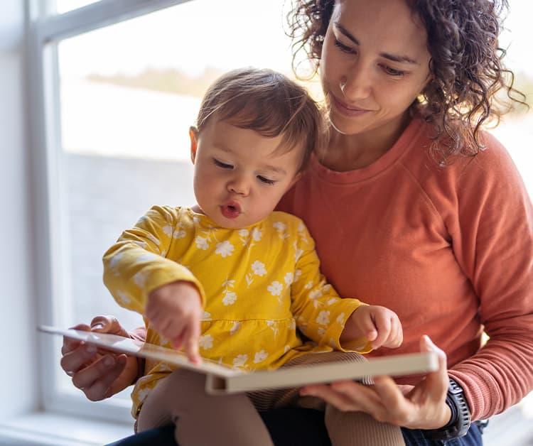 mother and child read a book together by a window. The child points at the book, wearing a yellow floral dress.